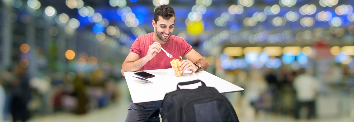 Een man in een rood T-shirt zit aan een tafel op het vliegveld. Hij lacht terwijl hij met een vork eet uit een fastfoodverpakking. Op de tafel liggen een smartphone en een instapkaart, en er staat een zwarte rugzak naast hem. De achtergrond toont een onscherpe, verlichte luchthavenomgeving.