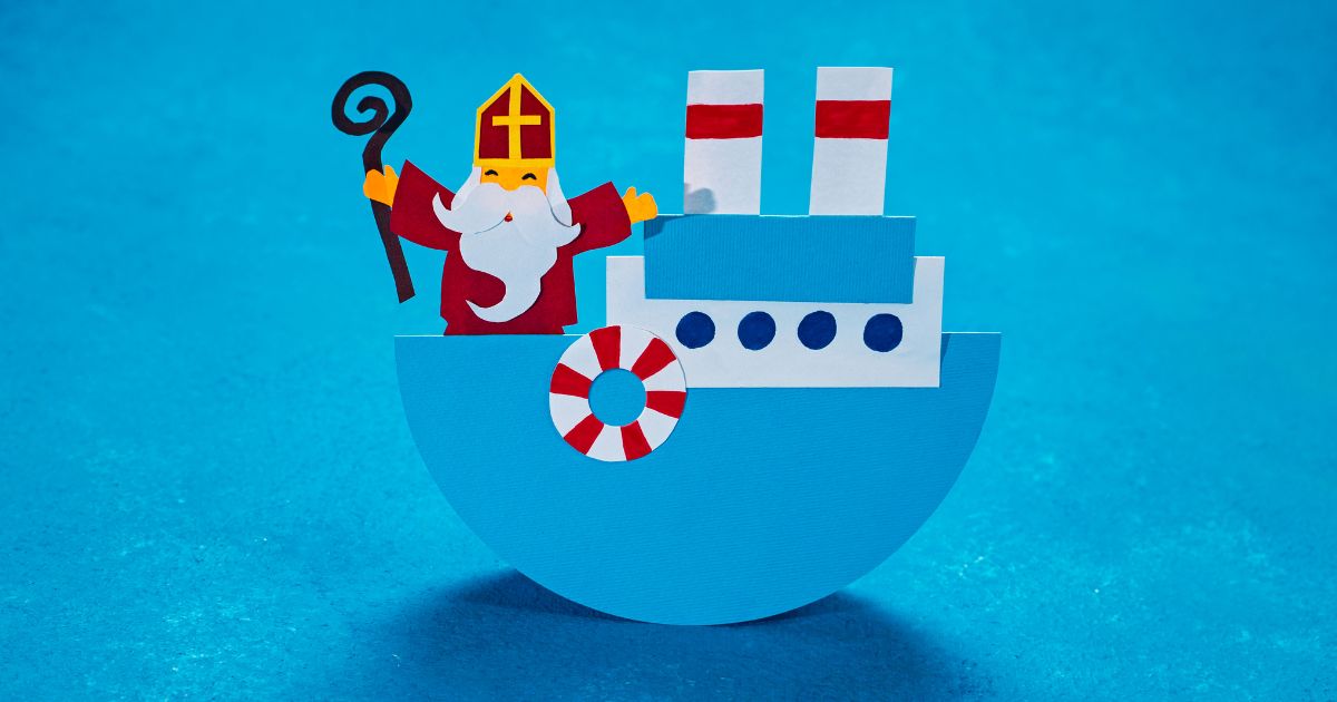 Papieren knutselwerk van Sinterklaas op een blauwe stoomboot, gemaakt van gekleurd papier. Sinterklaas draagt een rode mijter en houdt een staf vast, terwijl hij op de vrolijk versierde boot staat, met rode en witte schoorstenen en een reddingsboei.