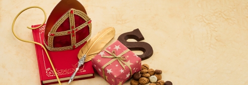 Sinterklaas stilleven met een rode mijter, een gouden staf, een boek met rode kaft, een veer, een ingepakt cadeautje, chocoladeletter 'S' en een hoopje pepernoten. De compositie symboliseert de traditionele Sinterklaasviering.